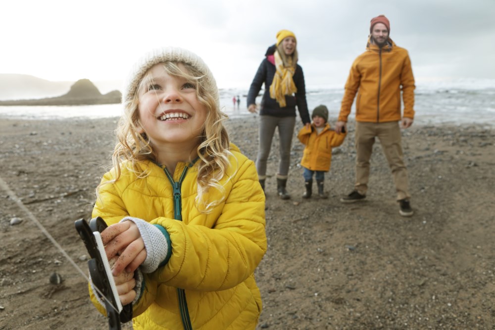 Smilende barn i gul jakke leker på stranden med foreldrene i bakgrunnen, som også har på seg gule jakker og ser på med glede.