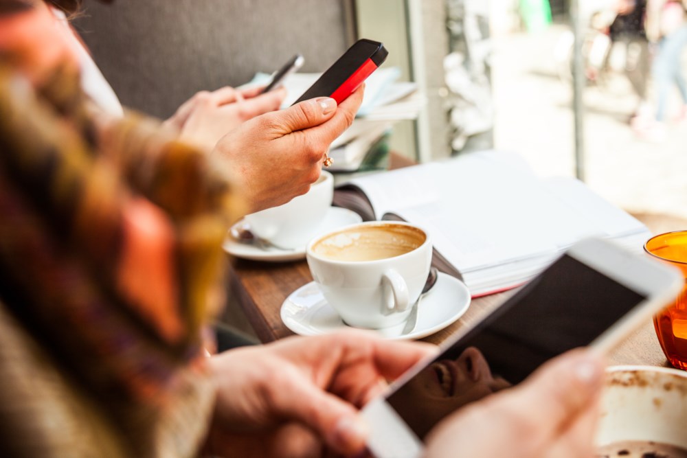 To personer ser på sine smarttelefoner mens de sitter ved et kafébord, symboliserer bruk av digitale betalingsløsninger i hverdagen.