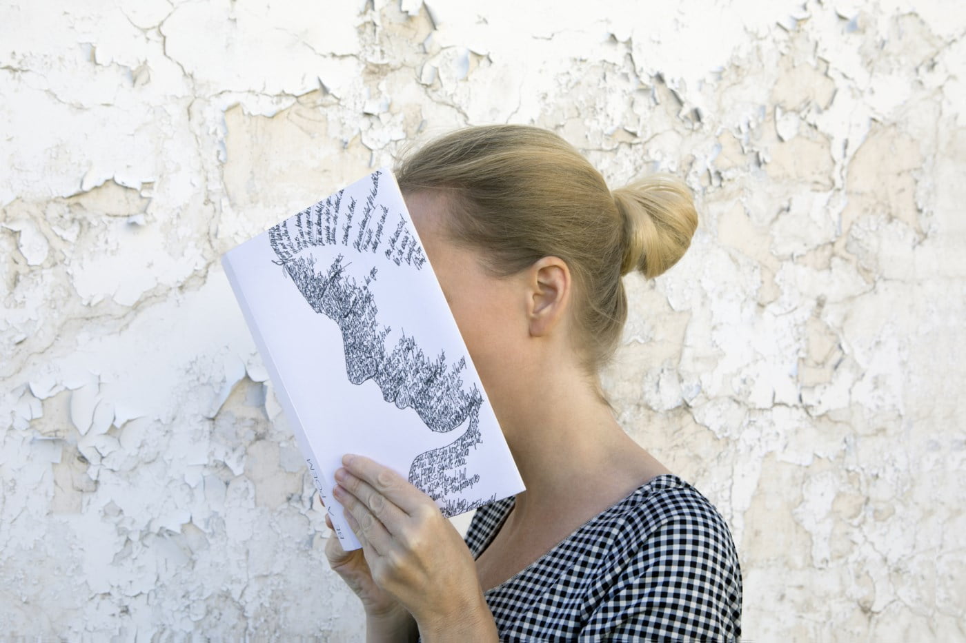 Kvinne holder en åpen bok foran ansiktet sitt, skjuler delvis sin identitet, mot en flassende vegg.