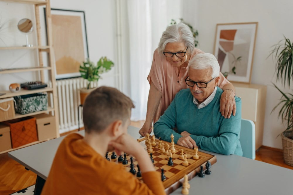 Besteforeldre spiller sjakk med barnebarnet sitt ved kjøkkenbordet, alle smilende og konsentrerte.
