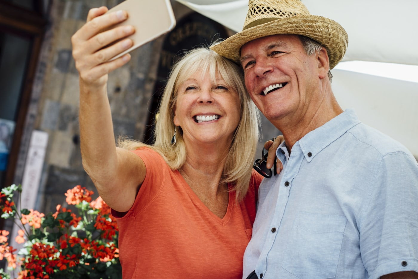 Eldre par tar en selfie mens de smiler bredt. Kvinnen i oransje t-skjorte og mannen i lyseblå skjorte, begge med sommerhatter, omgitt av blomster