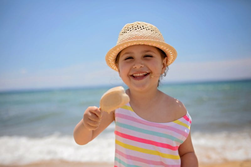 Liten jente i stripet kjole og stråhatt smiler mens hun holder en iskrem på stranden.