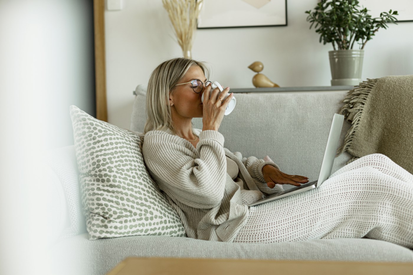 Kvinne lener seg tilbake på en sofa, drikker fra en kopp, og ser på en laptop på fanget sitt i et stilig innredet rom.