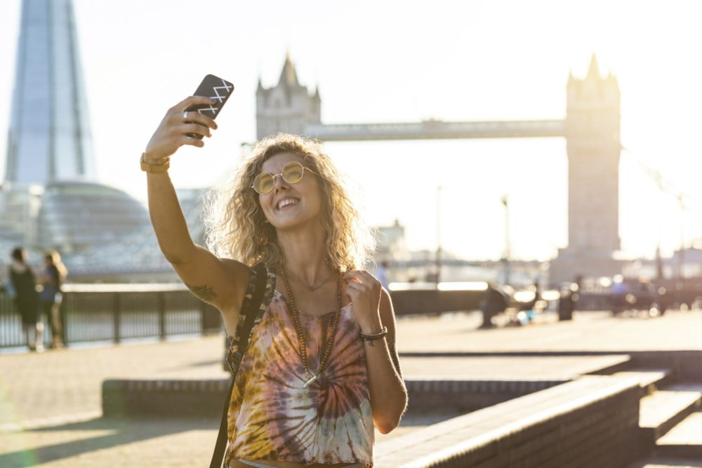 Kvinne på ferie tar en selfie foran Tower Bridge i London, smilende i solskinnet med solbriller og fargerik topp.