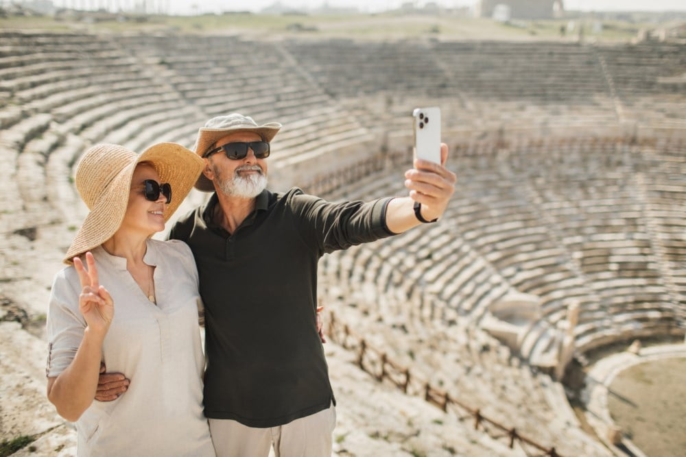 Eldre par tar en selfie foran et gammelt amfiteater, begge smilende med solhatter og solbriller.