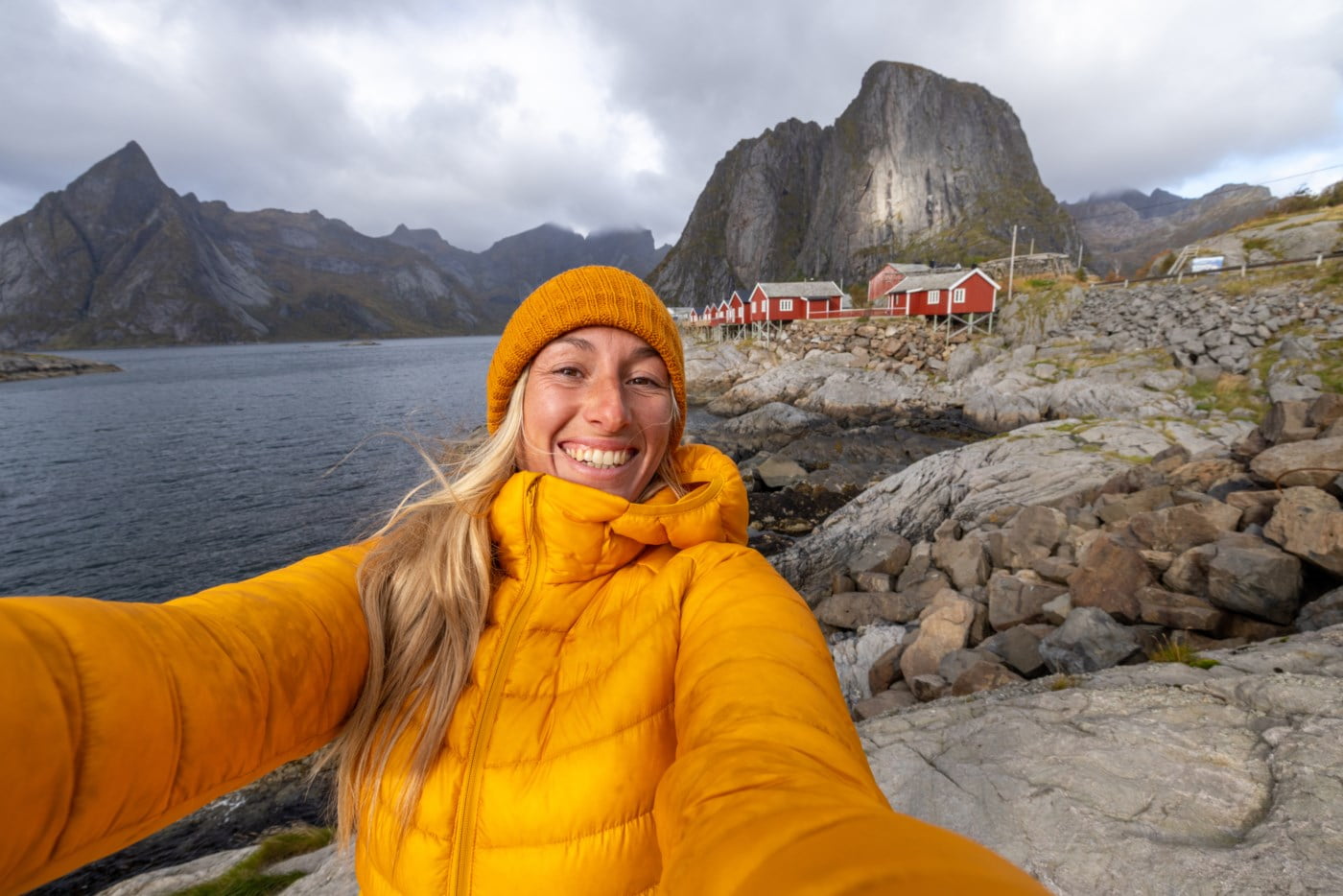 Strålende kvinne i gul jakke tar en selfie med den majestetiske norske kysten og røde hytter i bakgrunnen, illustrerer varme og glede i enkelheten ved livet.