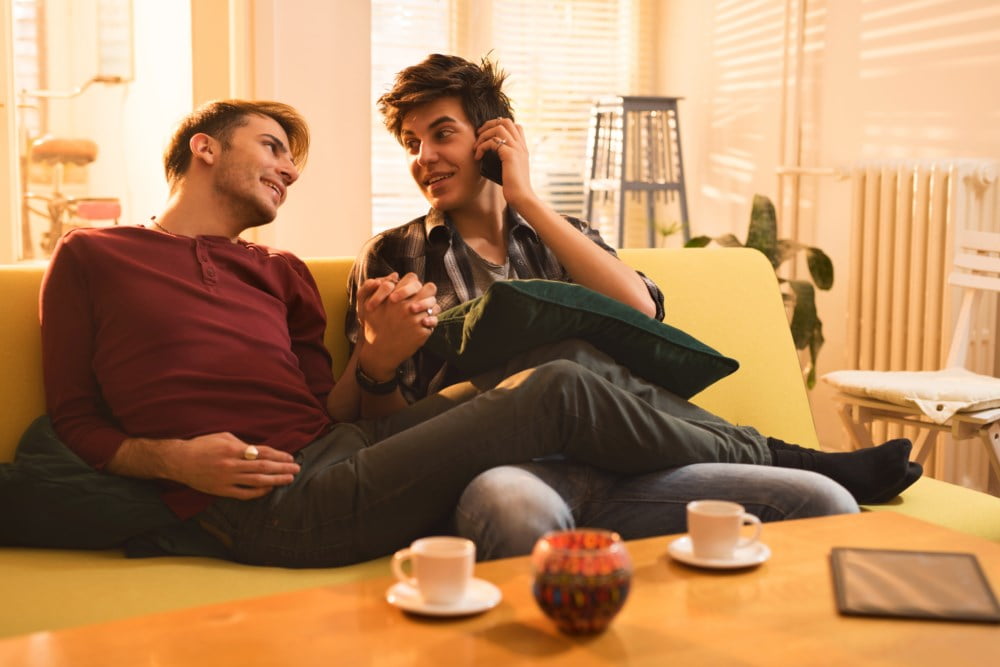 Et smilende par sitter på en sofa i sitt nye hjem, holder hender og snakker i telefonen.
