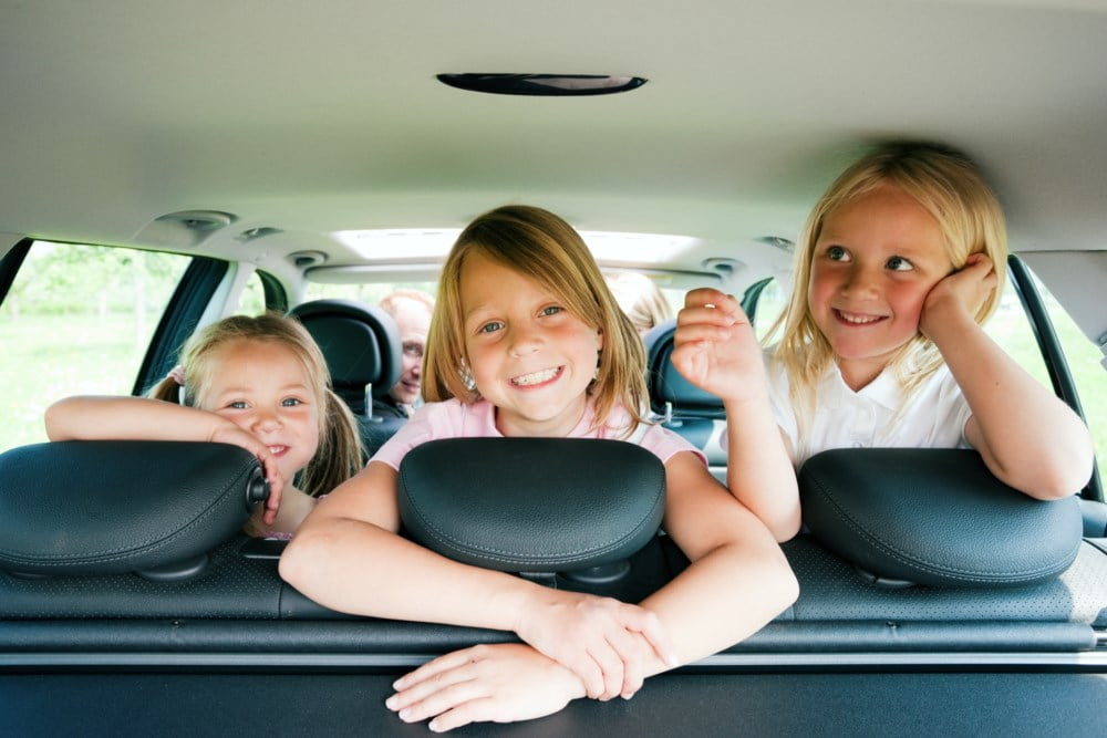 Tre glade barn sitter i baksetet på en bil, ser inn i kameraet og smiler under en familieutflukt.