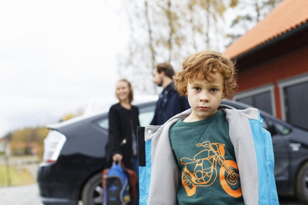 Liten gutt med ryggsekk ser tankefullt ut mens han står ved siden av en svart bil, med familie i bakgrunnen.