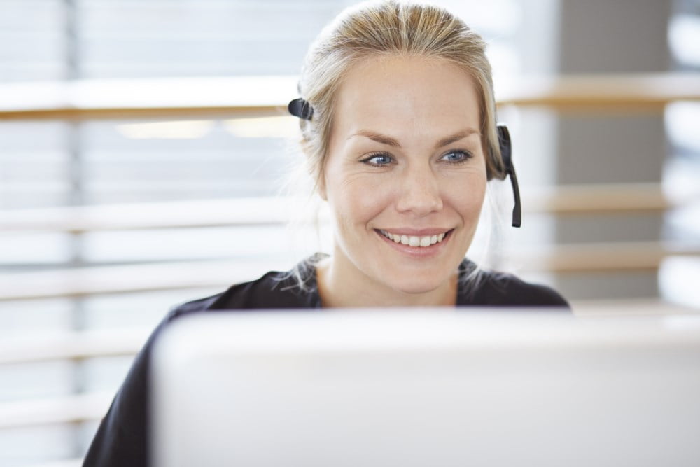 En blid kvinne med headset jobber på datamaskin, ser engasjert og lyttende ut i en lys og moderne kontorsetting.