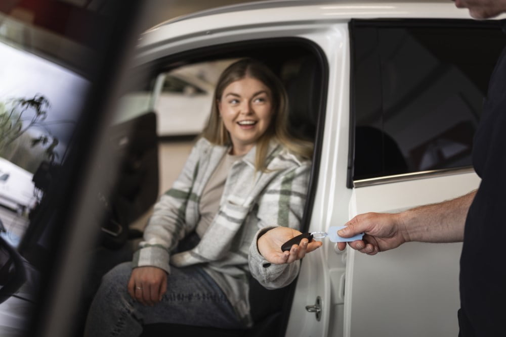En ung kvinne smiler bredt mens hun mottar bilnøkler fra en selger i en bilforretning.