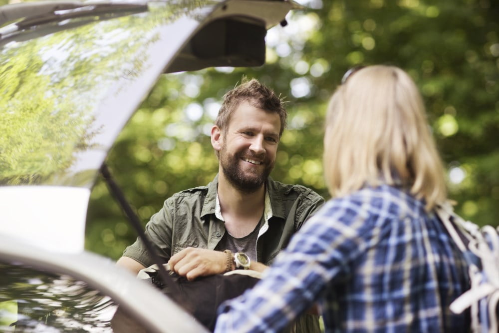 Smilende mann som lener seg på åpen bagasjeluken til en bil mens han snakker med en kvinne utenfor bildet, omgitt av frodig grøntområde.