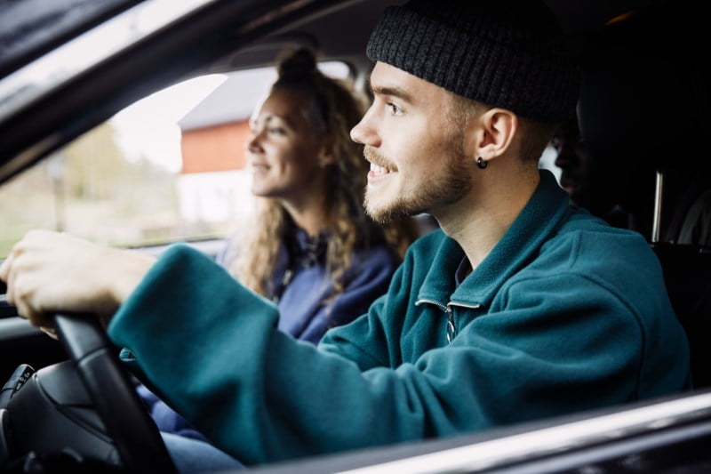 Et smilende ungt par i en bil, ser glad og avslappet ut mens de kjører gjennom et naturskjønt område.