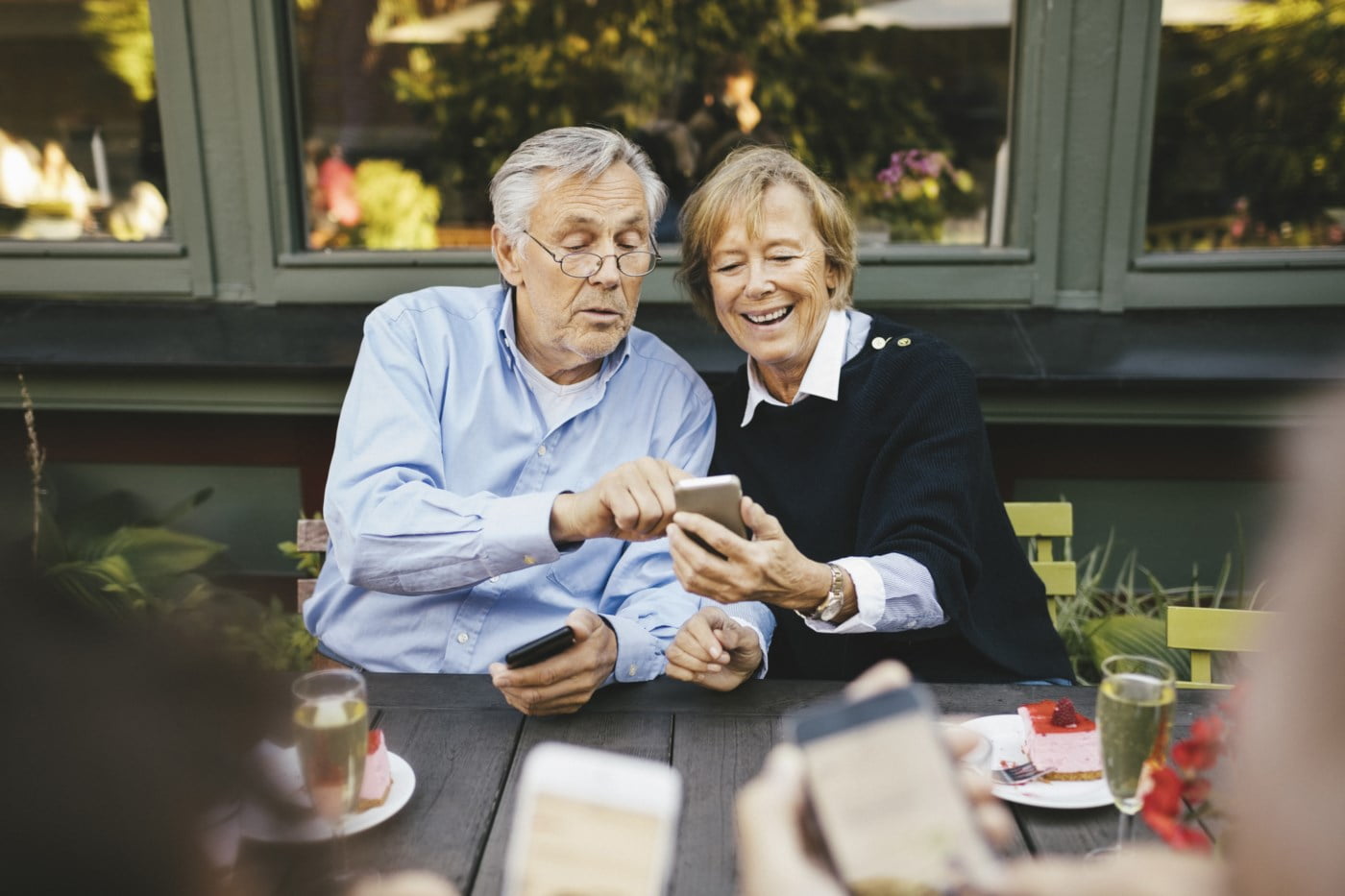 Et eldre par sitter ved et bord utendørs og ser på en smarttelefon sammen, begge smiler og nyter øyeblikket.
