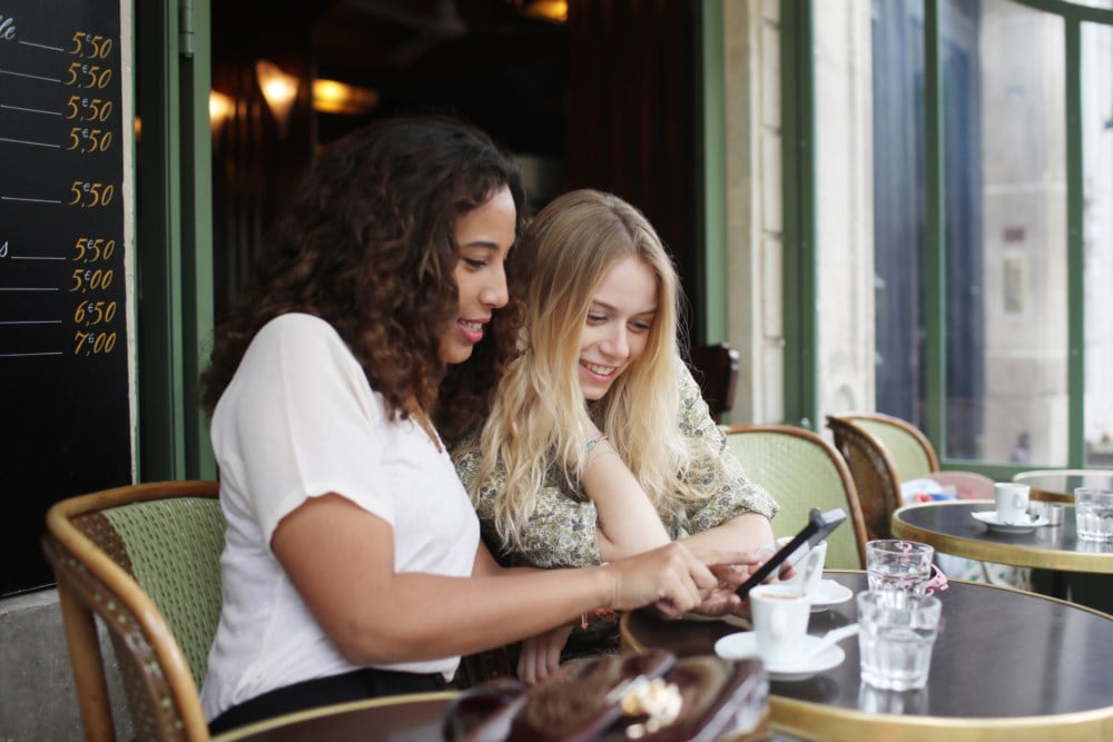 To unge kvinner sitter ved et kafébord, smiler og ser på en mobiltelefon sammen.
